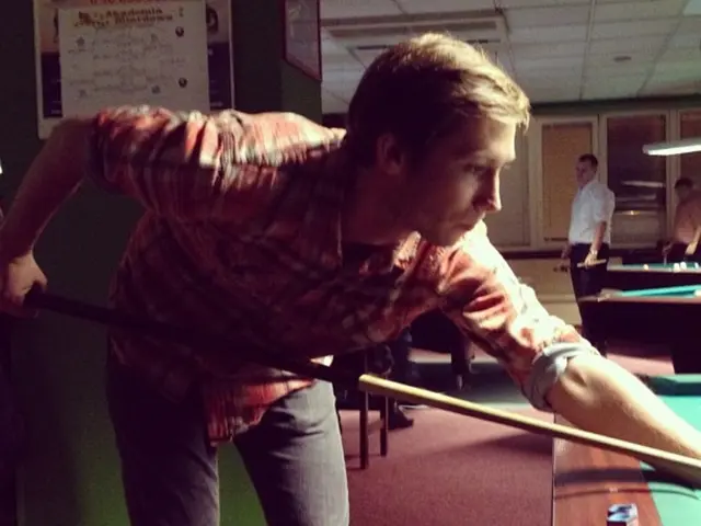 The man in red check shirt and the black pant is playing a snooker pool game. Beside him, we see...